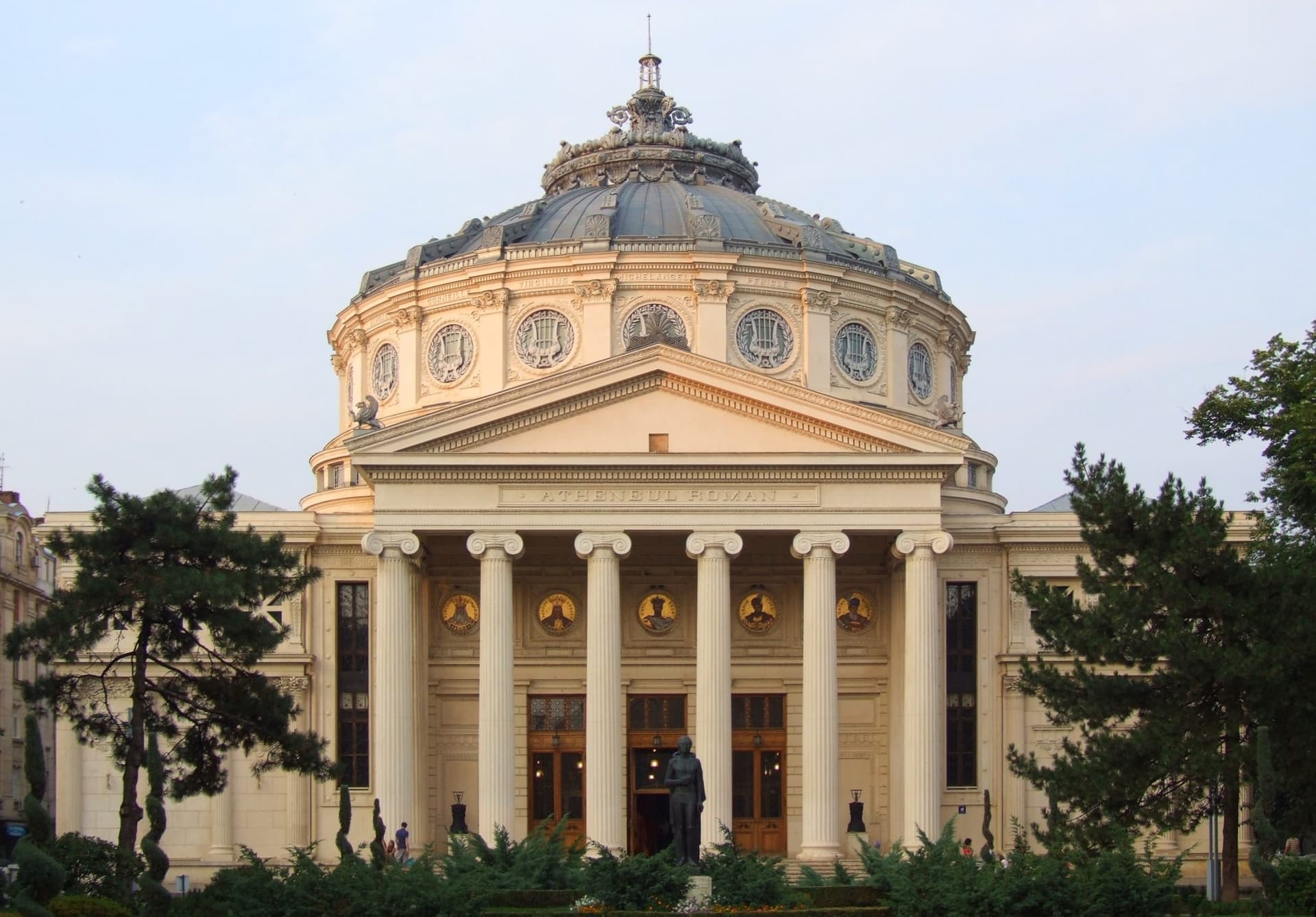 Fațada Ateneului Român din București în lumina serii: peristilul cu șase coloane ionice, frontonul cu inscripția și rotonda cu cupolă, cu statuia lui Mihai Eminescu în fața intrării și brazi de o parte și de alta.