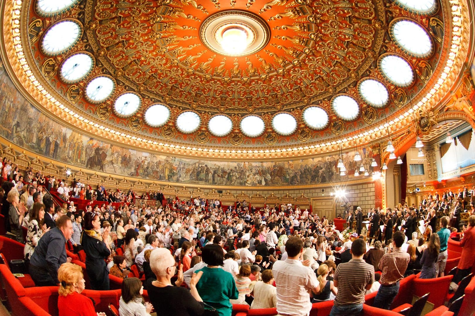 Sala mare a Ateneului Român, fotografiată de sus: cupola aurită cu fresca circulară, lojele roșii și publicul care aplaudă în picioare.