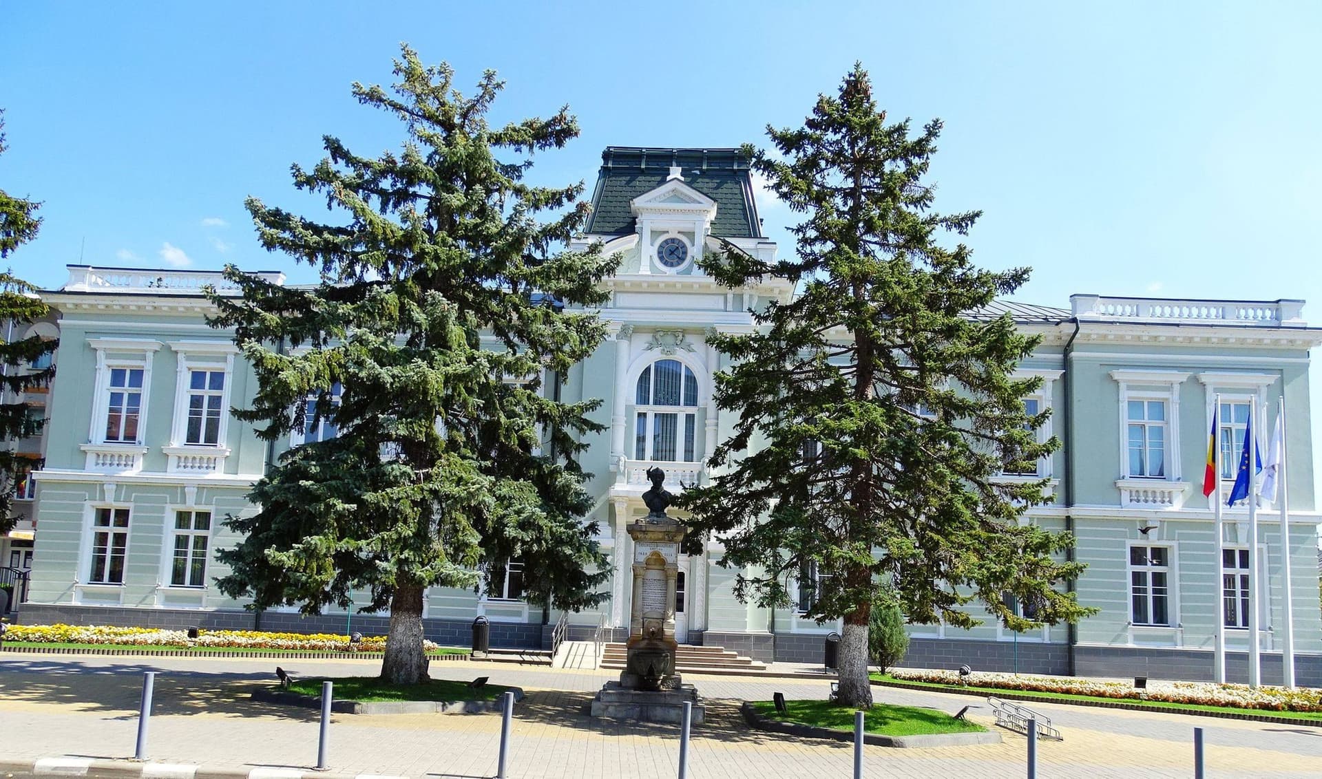 Fațada principală a clădirii Primăriei Râmnicu Vâlcea, monument istoric din 1912, cu ceas deasupra intrării, doi brazi înalți în fața ei și drapelele României și Uniunii Europene arborate pe latura dreaptă.