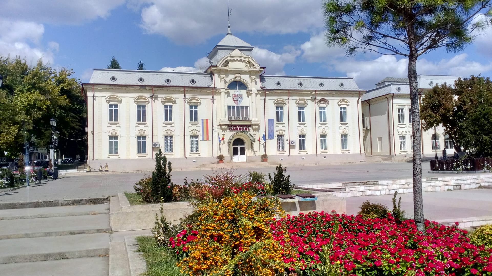 Clădirea Primăriei municipiului Caracal, un edificiu alb în stil eclectic, cu turn central cu ceas și cupolă, steaguri la intrare și un rond de flori roșii și galbene în prim-plan.