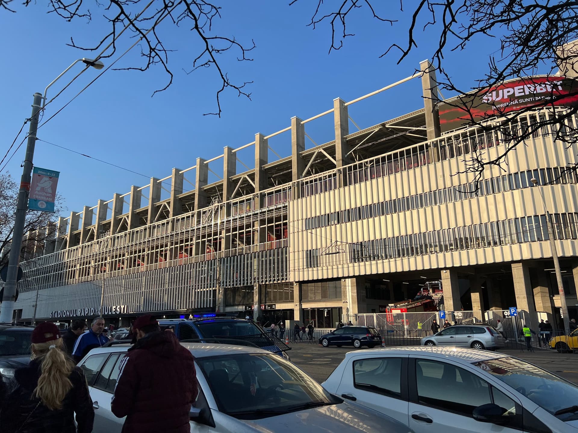Exteriorul stadionului Rapid-Giulești din București, cu fațada iluminată și fani adunați în fața intrărilor, seara.
