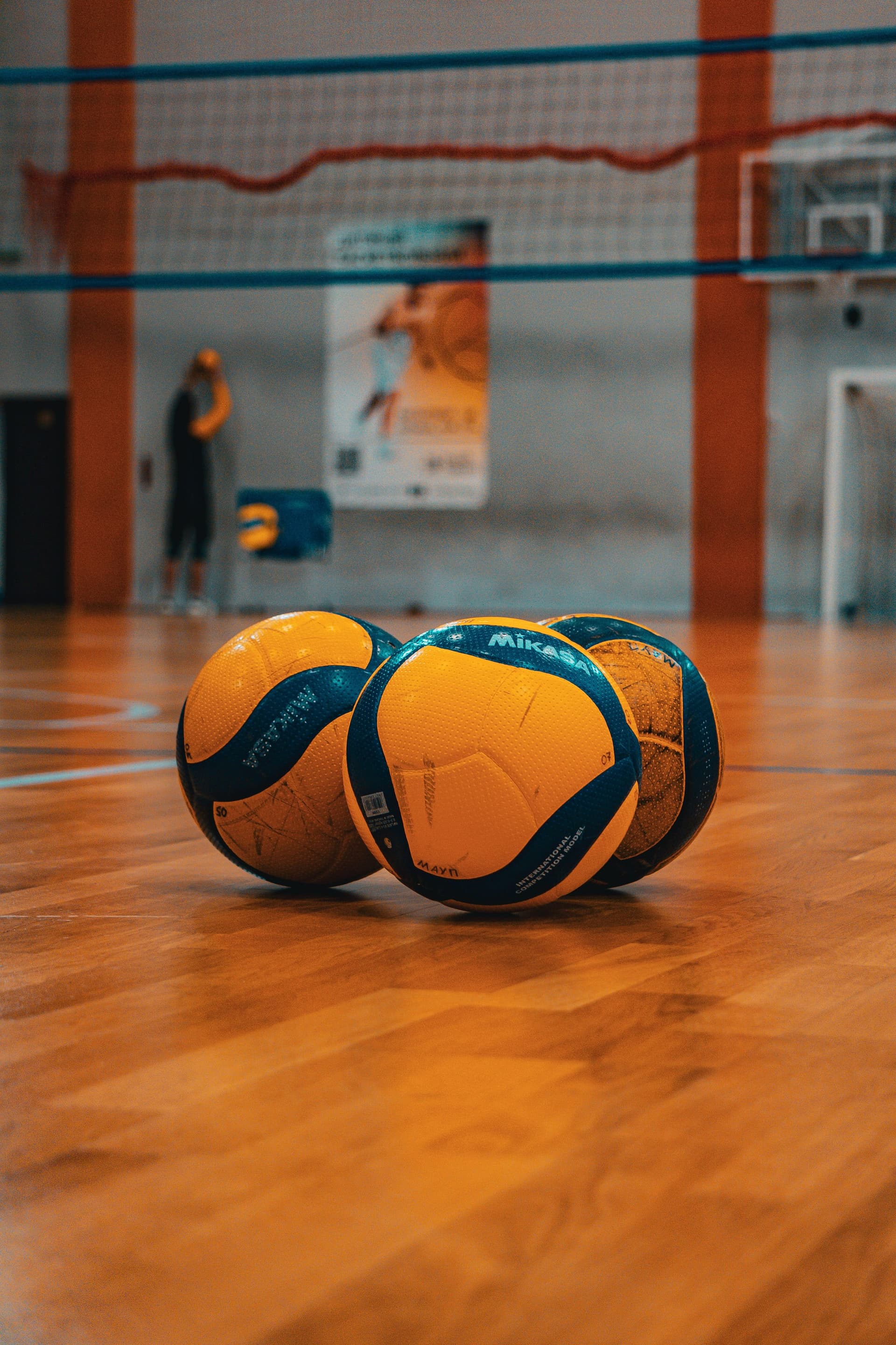Trei mingi de volei galben-albastre așezate pe parchetul lustruit al unei săli de sport, cu fileul de volei întins în plan secund și o siluetă neclară de jucător în fundal.