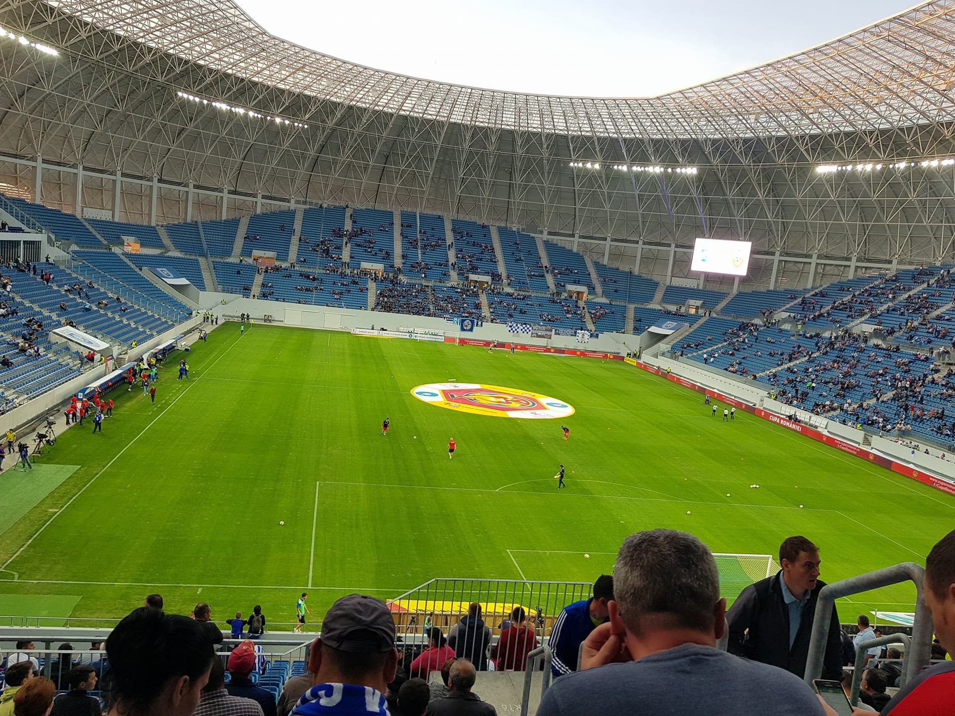 Interiorul stadionului Ion Oblemenco din Craiova, văzut din tribună: gazon verde, emblema Universității Craiova la centrul terenului și scaune albastre sub acoperișul alb.
