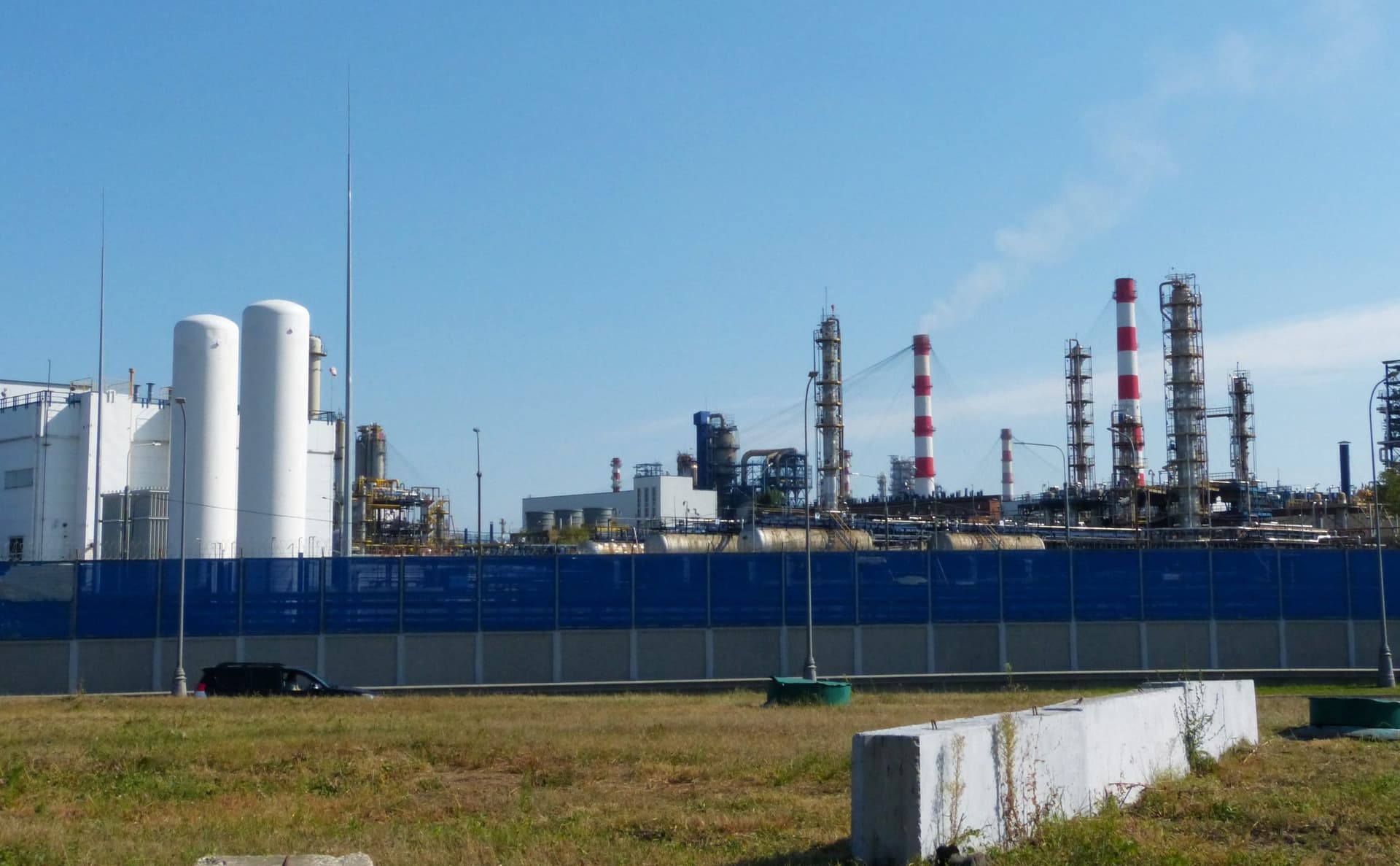 Vedere de la nivelul solului către rafinăria de petrol din Moscova, în cartierul Kapotnia: rezervoare albe cilindrice, coloane de distilare metalice și coșuri vopsite în dungi roșii și albe, în spatele unui gard albastru înalt, sub cer senin.