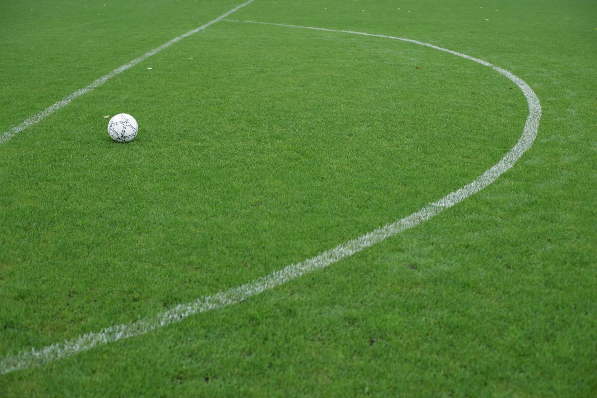 Minge de fotbal albă așezată pe gazonul verde al unui teren de fotbal, lângă linia albă curbă a cercului de la centru.